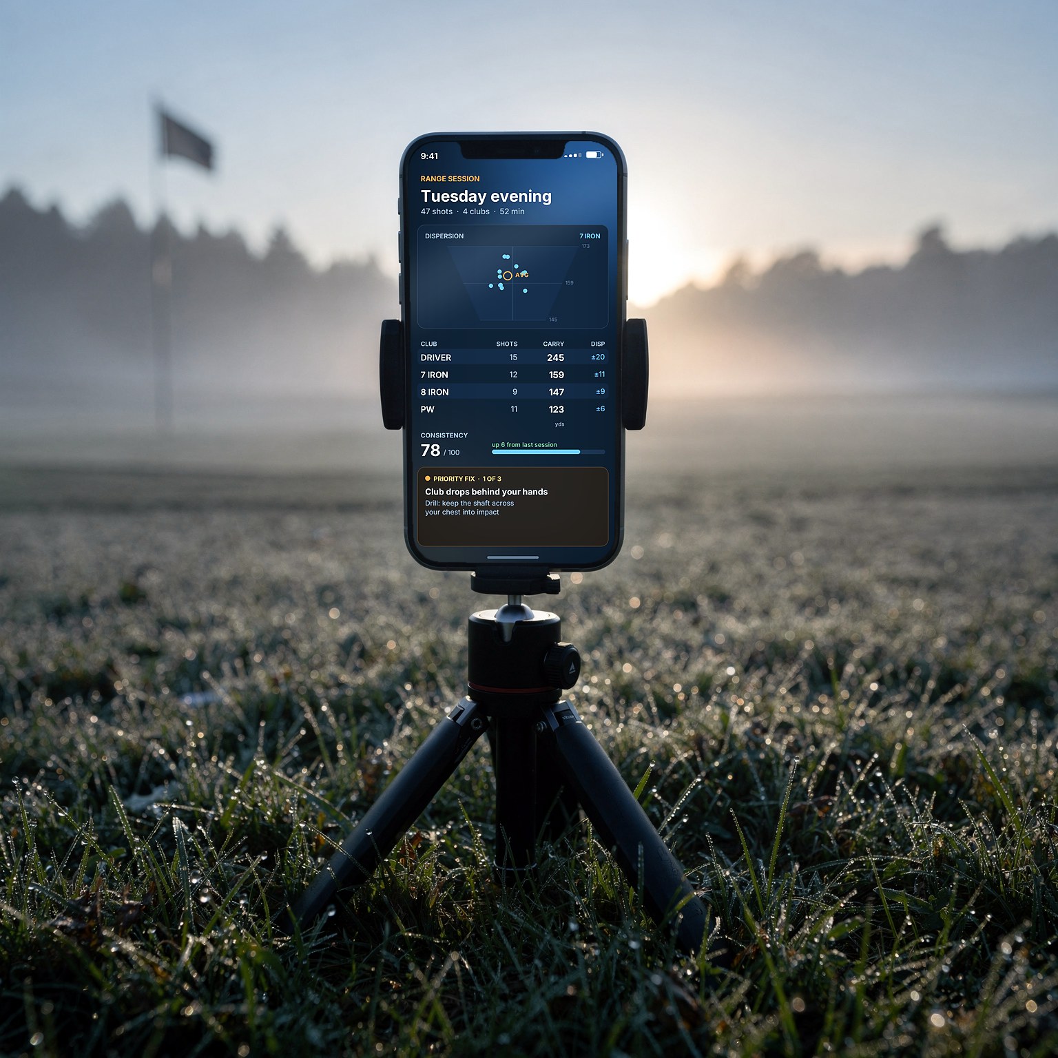 A phone standing upright on a small tripod at a misty dawn driving range, its screen showing a Golfer Awakens range session summary: shot dispersion, carry distances by club, a consistency score and the priority fix